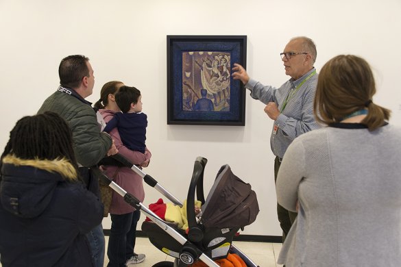 Several people, including one woman holding a small child, gather near a docent talking in front of a painting of several can-can dancers