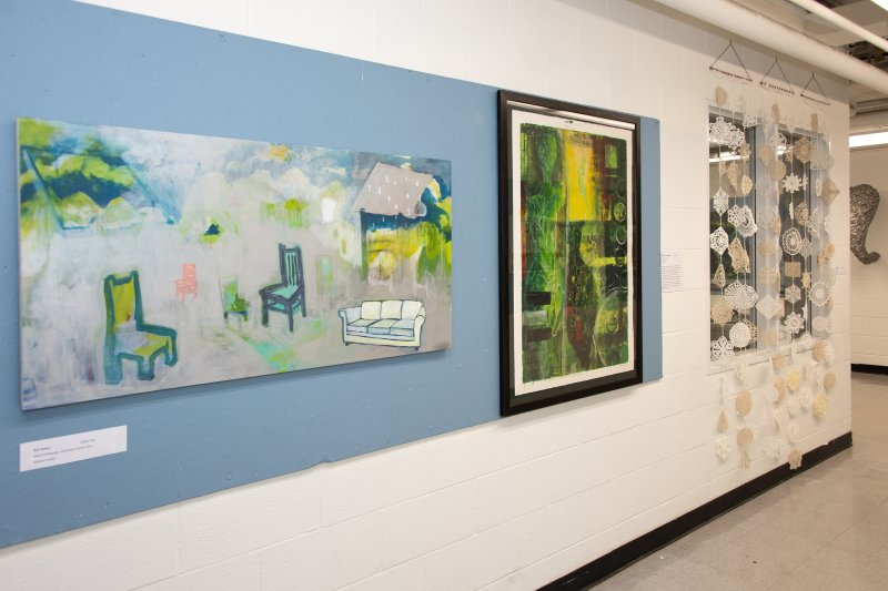 A painting of chairs, an abstract painting with yellows and greens, and delicate paper sculptures hanging over a window