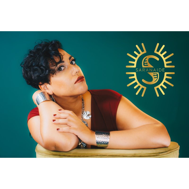 A woman posing with her head resting on her arms with a green background and a gold emblem that says "Saranaide"