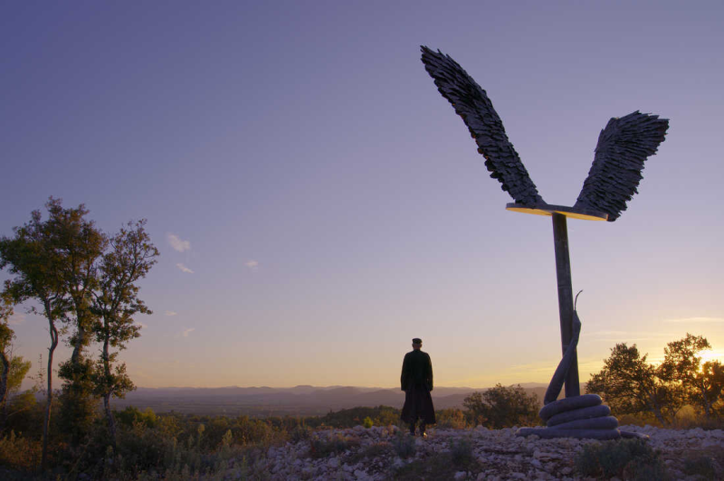 Film still of a person's silhouette next to a large statue of wings outside at sunset