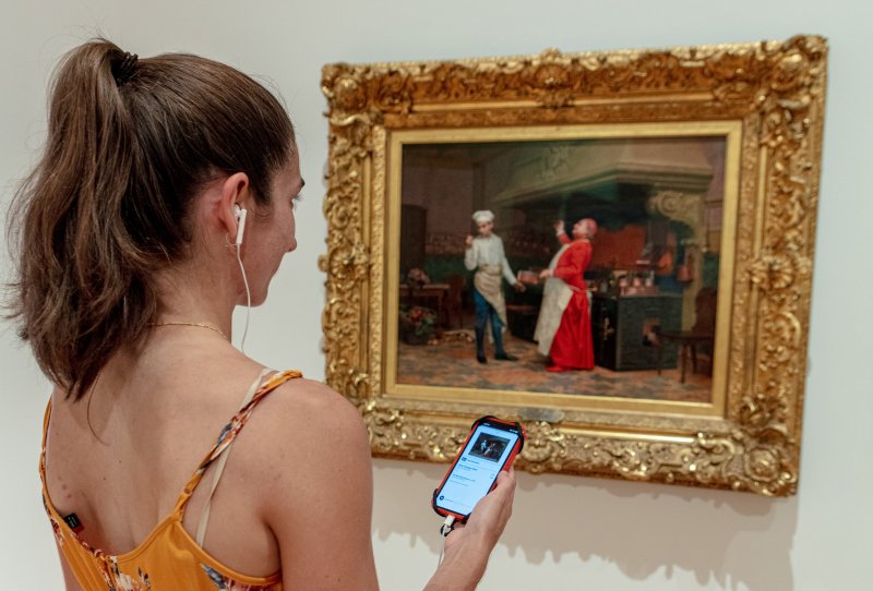 A woman with headphones and a cellphone looking at a painting