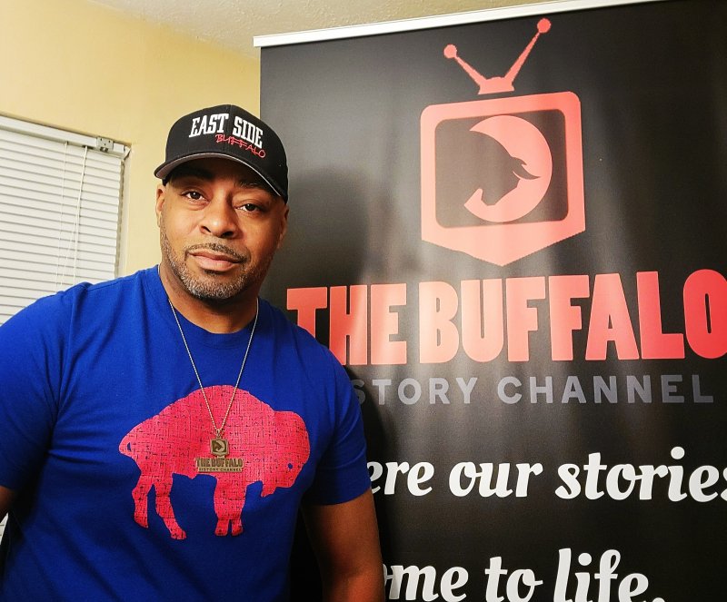 A man with medium-dark skin tone wearing a black baseball hat and blue shirt with a red buffalo on it