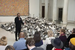 An Indian man stands in front of a group of people sitting on chairs, next to his work which is made up of hundreds of metal pots and pans