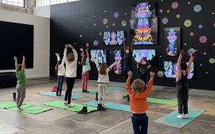 Kids doing yoga in front of an artwork made of several TV screens hanging on a black wall showing a colorful being