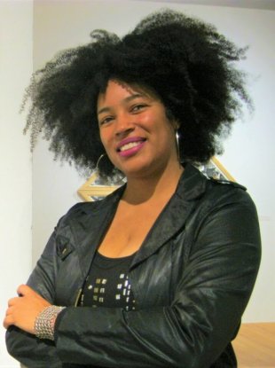 A Black woman in a black leather jacket, seen from the waist up, stands with arms cross, head cocked to one side