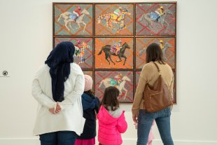 Two women and three kids looking at an artwork of nine panels with jockeys on horses in various colors