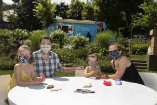 A man, a woman, and two kids wearing face masks sitting at a table in a garden with the Art Truck parked on the street in the distance behind them