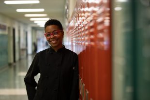 An African American woman in a black chef smock and red glasses stands in front of a bank of red lockers