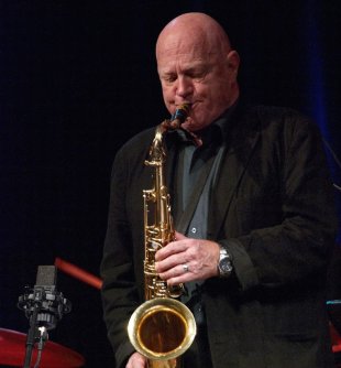 A bald man in a dark suit playing the saxophone