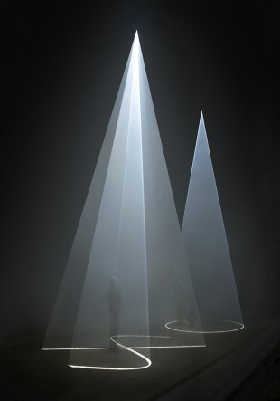 A photograph of three people standing in a dark room, engulfed in two cones of bright white light projected from the ceiling onto the floor