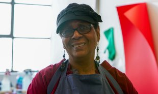 A Black woman with a black hat, glasses, and long beaded earrings wearing a dark red shirt and blue apron
