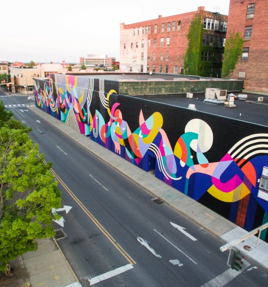 From a treetop view, an energetic patterning of abstract shapes in vibrant hues of magenta, salmon, teal, navy, mustard yellow, and tomato red covers the façade of a single-story building that stretches the length of a city block. These forms dance along a black background overlaid at point with black and white striped designs.