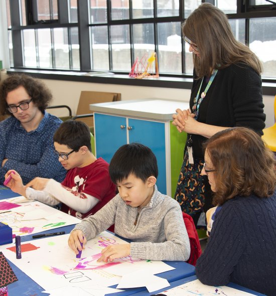 Two adults sitting next to two kids, with one adult behind them to guide the kids&#039; artmaking