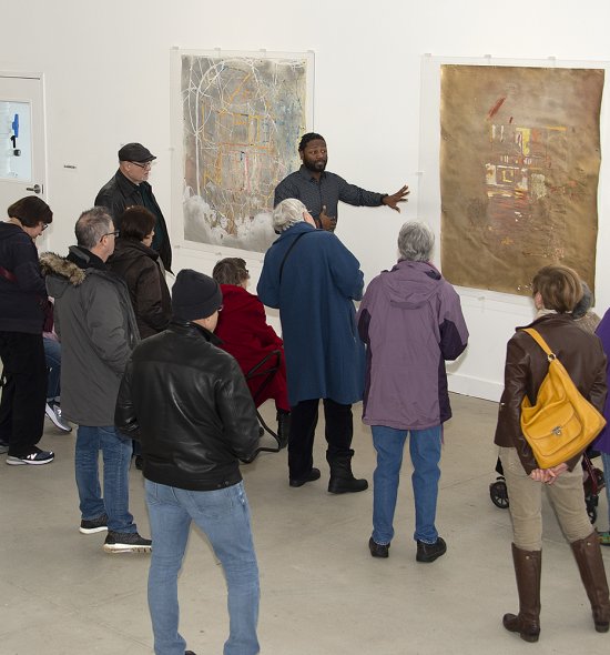 An African American man talks to a group of people while in front of a large gold-colored painting
