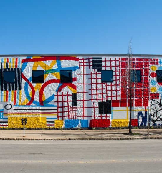 A mural on a long exterior wall featuring different painted sections using red, yellow, and blue to create different patterns