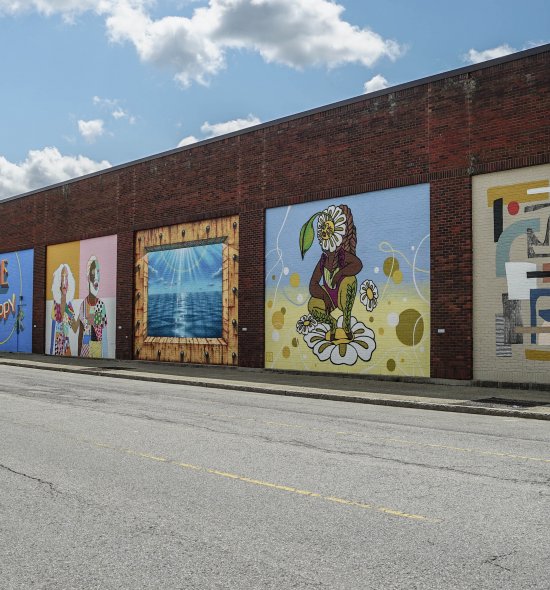 A receding line of varied murals on a long brick wall with blue, cloudy sky above.