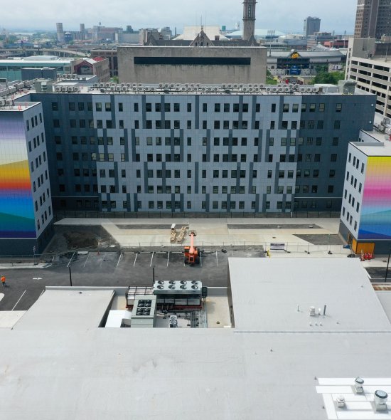 An elevated photograph of Josef Kristofoletti&#039;s Golden Hour, a mural that covers four faces of the building at 201 Ellicott Street in bands of bright color. In the background is the cityscape of Buffalo.