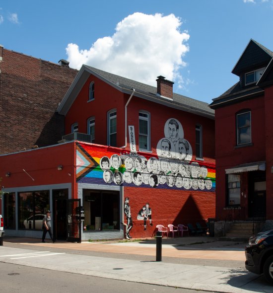 Mural of the faces of LGBTQ+ icons against the Progress Pride flag on a red brick building with city street and blue sky in background.