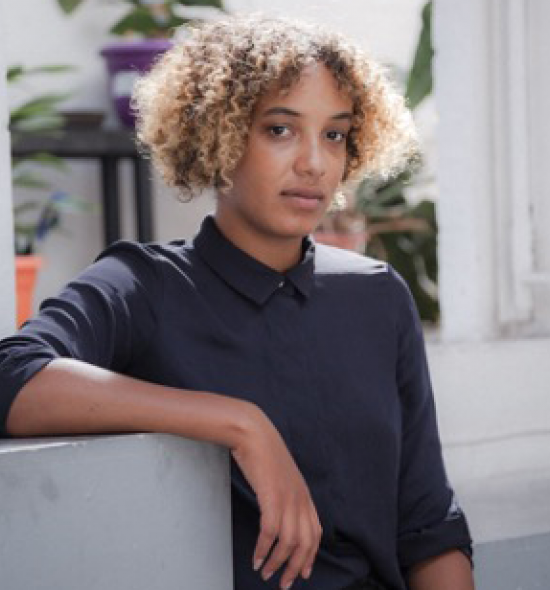 Woman of medium-light skin tone and curly blonde hair in black shirt rests right arm on a shelf, white room with plants in the background