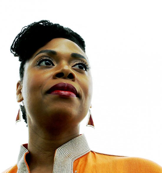 A woman of dark skin tone seen from the shoulders up, in an yellow blazer, with red earrings, and her hair in twists, photographed from below and looking up and to the right.