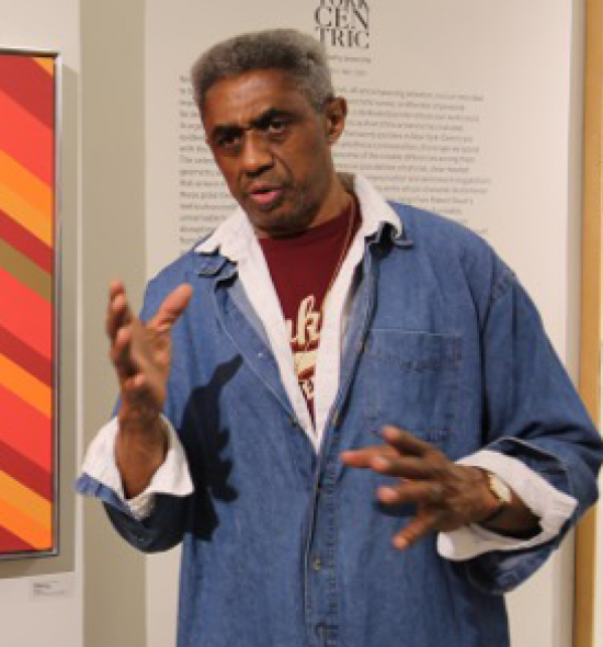 Man of dark skin tone in blue denim shirt speaking, hand gesturing in the air as if grabbing, at his left a painting of brightly colored diagonal stripes, predominantly orange
