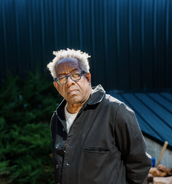 Photo from the waist up of a man of medium dark skin tone with gray hair and glasses, and black coveralls, behind a blue roof and siding and green shrubbery