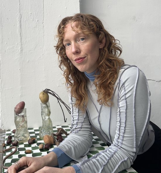 A woman with red wavy hair, fair skin with freckles, wearing a blue shirt leaning on a checkerboard table with potatoes sprouting in various bottles