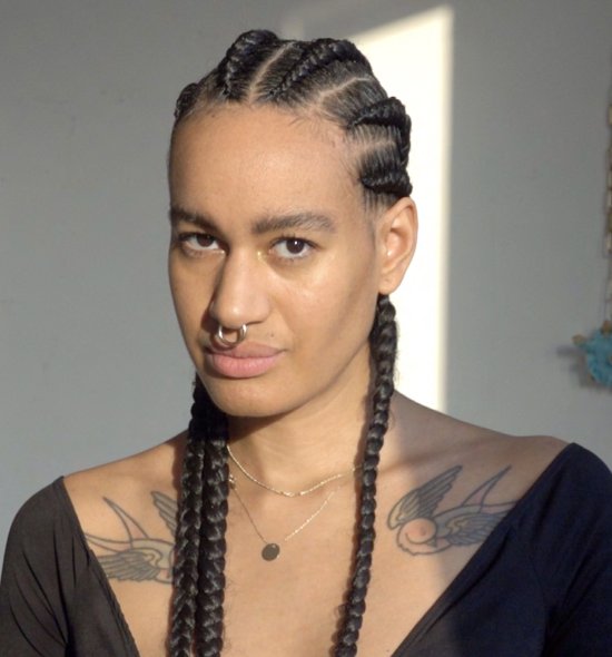 Headshot of a person with long dark braids, medium skin tone, and tattoos on their collarbones wearing a black v-neck sweater