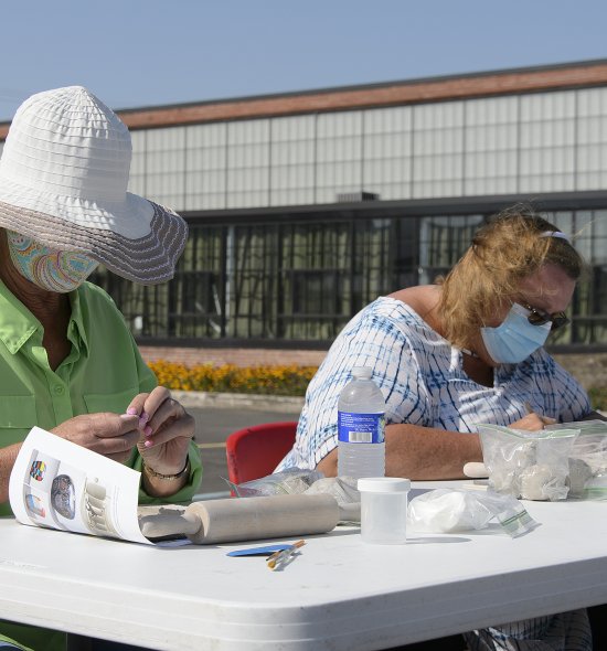 Two white women wearing face masks seated at a table making ceramic beads