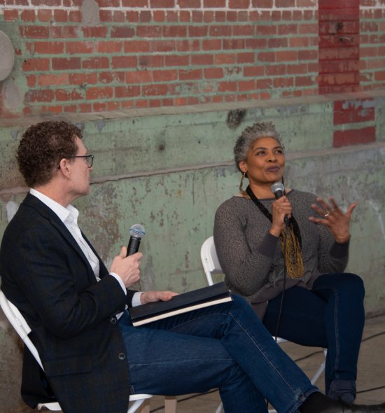 An African American woman talks into a microphone while a white man looks on