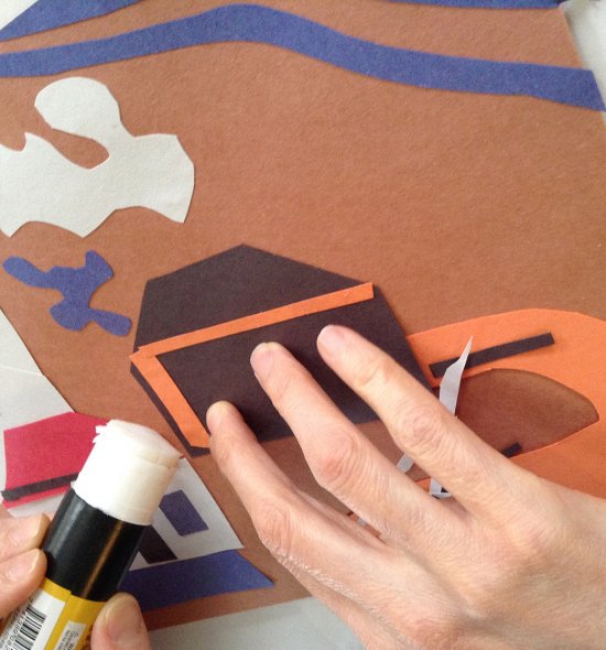 A hand gluing paper shapes to a sheet of brown paper