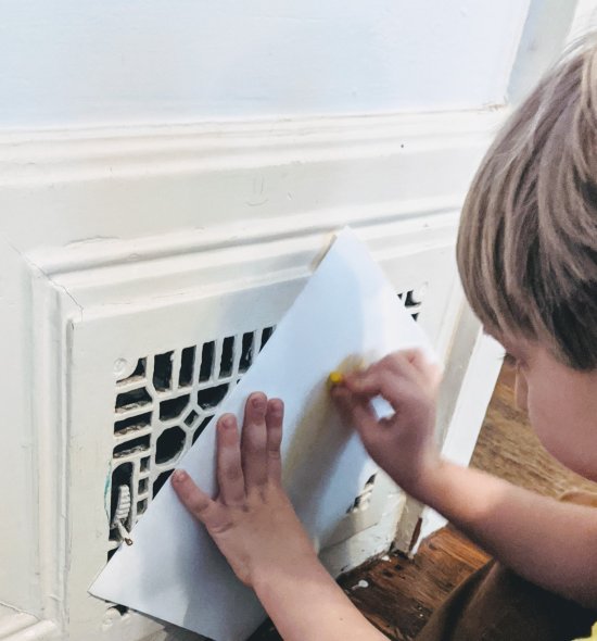 A boy holding a paper over a heating vent and rubbing a crayon on it