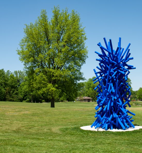 A large sculpture made out of long wooden poles painted bright blue arranged in a tower. The sculpture is set in the middle of a park with green grass and a large tree.