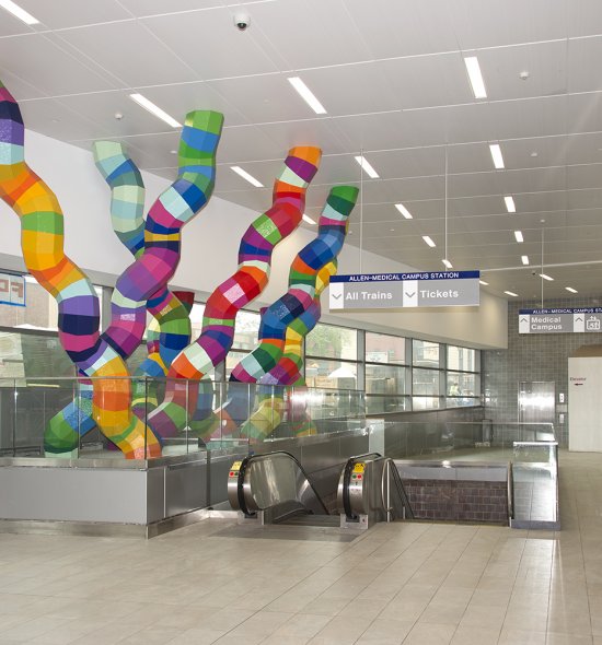 Six massive twisting columns segmented in bright shades of pink, purple, yellow, orange, green, and blue span from floor to ceiling slightly to the left of center in this image. These are shown installed in the brightly lit, largely white and light gray entryway to a subway station; directional signs hang from the ceiling, and the tops of a pair of descending escalators are visible to the right of the sculptures.