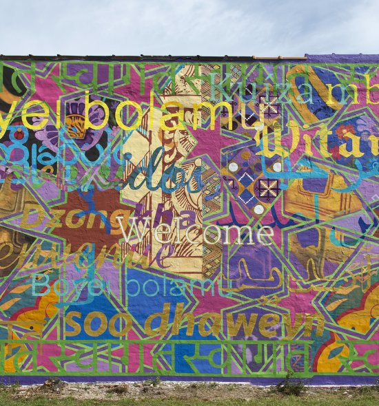 This brightly colored mural features the word “welcome” in thirteen different languages atop a dazzling background of magenta stars interspersed among geometric shapes in solid shades of blue, purple, and brown and graphically patterned areas.