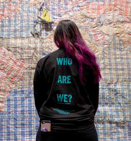 Woman with purple and black hair in a black long-sleeve t-shirt that says &quot;Who Are We?&quot; on the back
